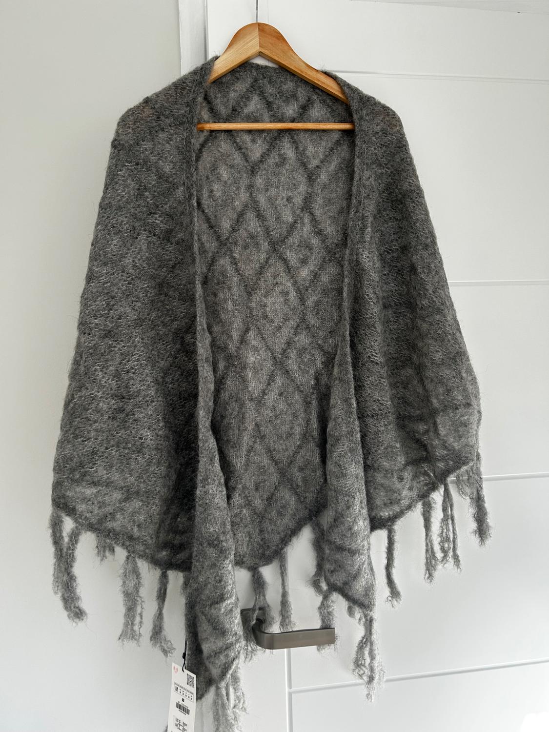 Grey fringed shawl hanging on a wooden hanger over a white door. Label visible in the bottom left.