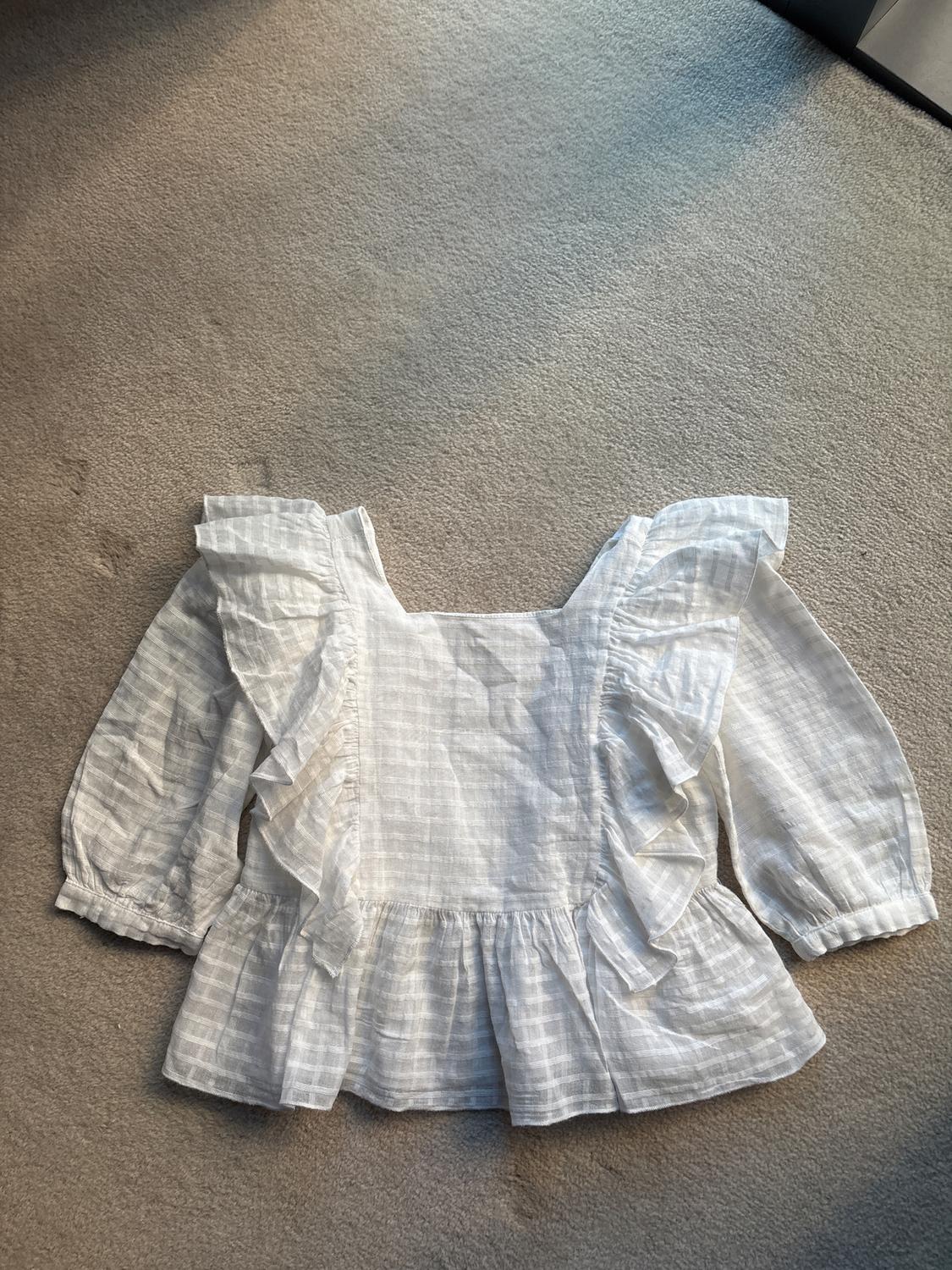Long-sleeved white blouse with ruffles and a checked design on a beige carpet illuminated by natural light.