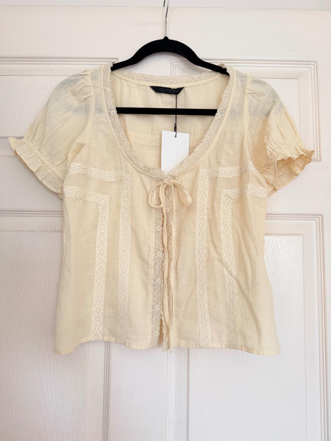 Cream short-sleeve blouse with lace and a front bow, hanging on a hanger over a white door. Text: ZARA.