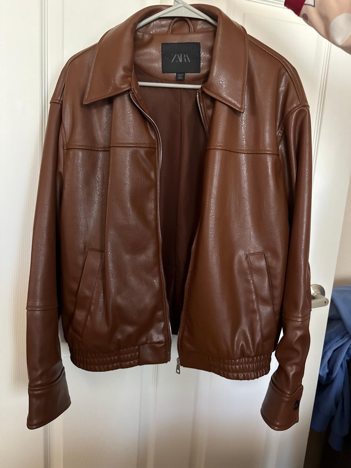 Brown faux leather jacket hanging on a hanger, with a front zip and a visible Zara label on the collar.
