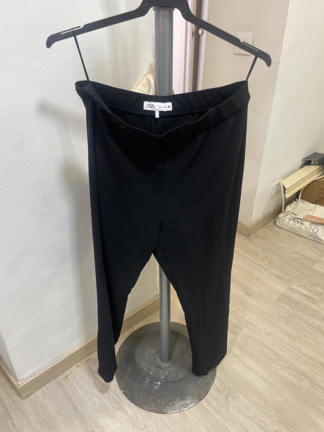 Black trousers hanging on a hanger on a metal stand in a room with a wooden floor. Label visible.