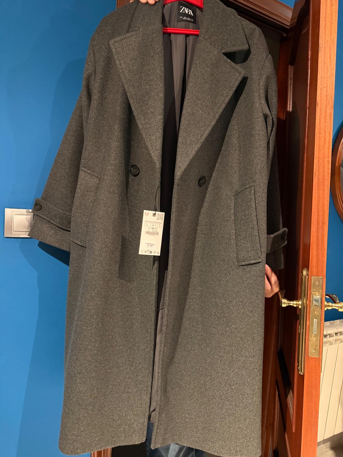Grey wool coat hanging on a red hanger, with a visible tag. Blue background and wooden door.