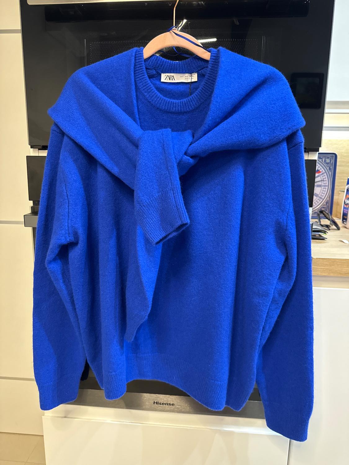 Blue jumper hanging on a hanger, with another blue jumper tied over the shoulders. Kitchen background with visible appliances.