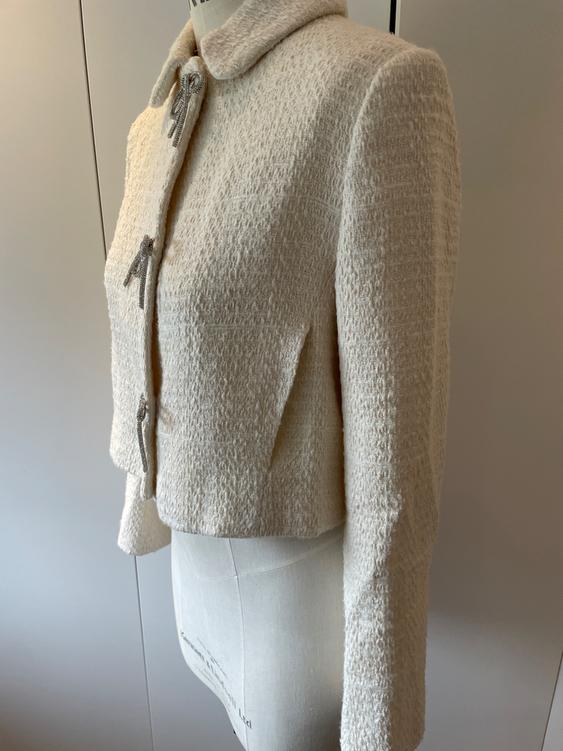 Thick-textured short cream jacket on a mannequin, with a decorative bow at the neck and a neutral background.
