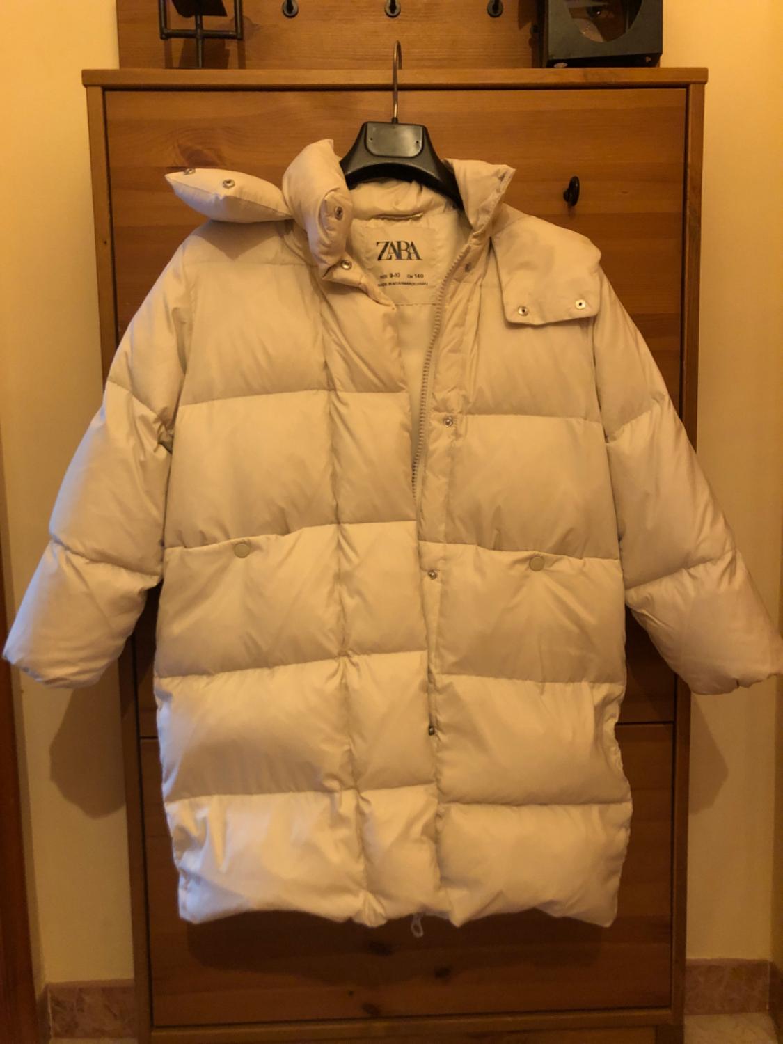 Beige padded coat hanging on a hanger in front of a wooden wardrobe. The label says "ZARA".