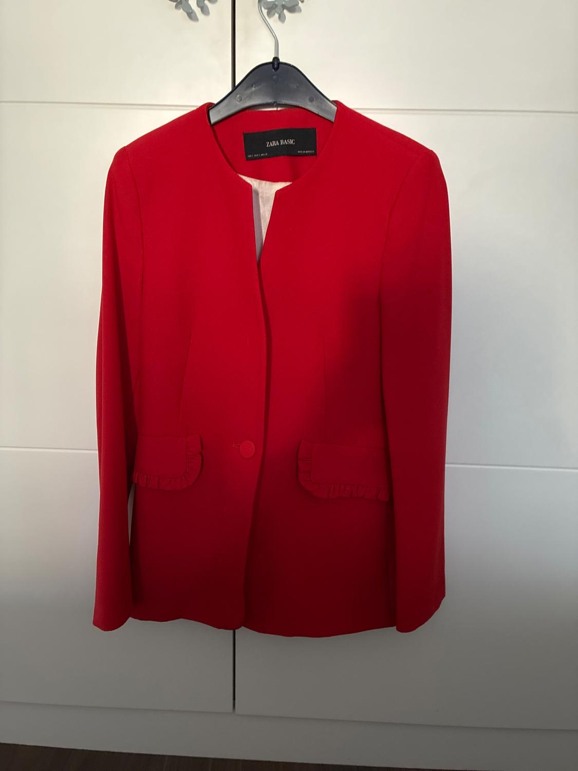 Red jacket hanging on a black hanger in front of a white wardrobe. It has one button and pockets with ruffles. Text: ZARA BASIC.