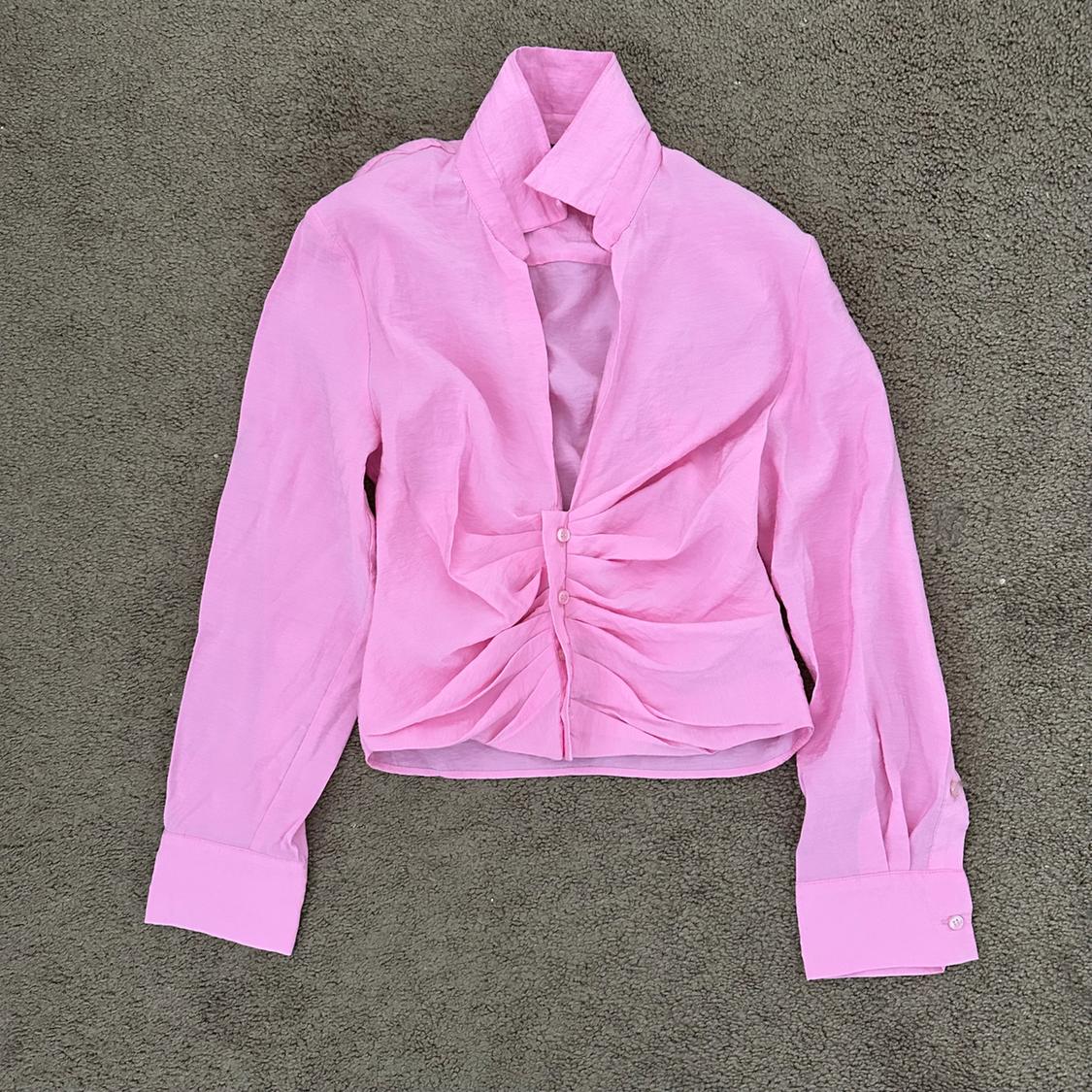 Pink long-sleeved blouse with front pleats and a high collar, on a gray rug.