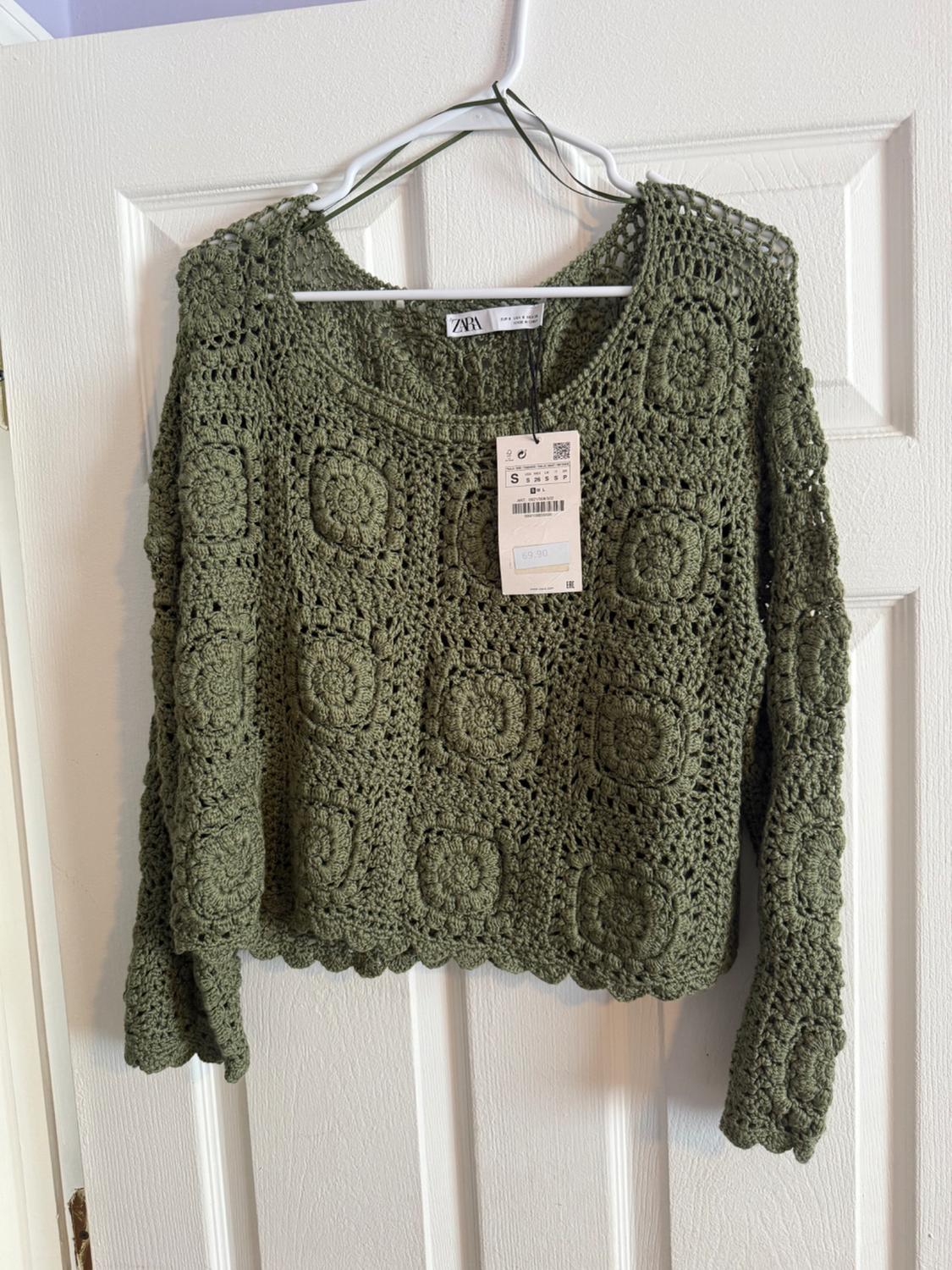 Olive green crochet jumper hanging on a white hanger over a white door. Label visible with the Zara logo.
