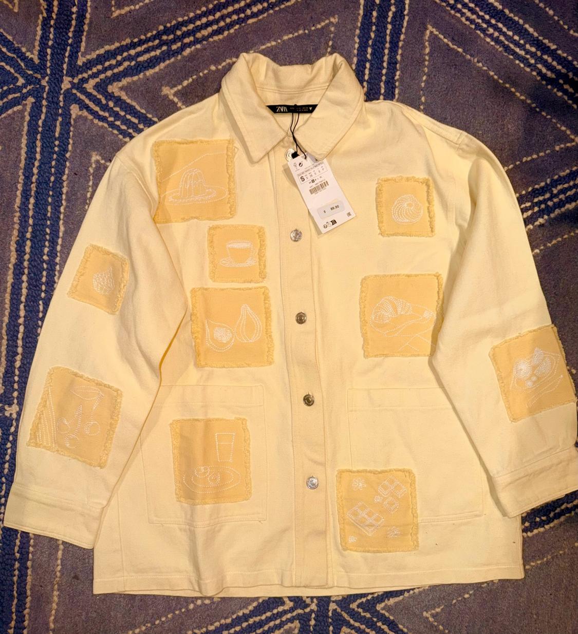 Yellow jacket with frayed patches and white embroidery, on a blue and white rug. Label with ZARA text.
