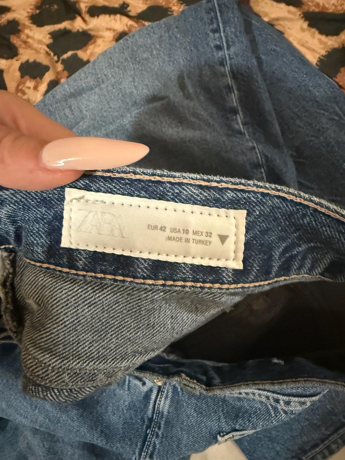 Close-up of a hand holding blue jeans. The label reads: ZARA EUR 42 USA 10 MEX 32 MADE IN TURKEY. Leopard-print background.