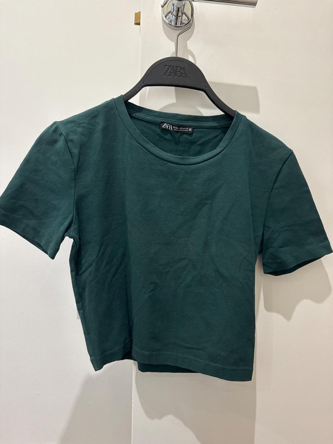 Green short-sleeved t-shirt hanging on a black hanger with the brand visible, against a white background.