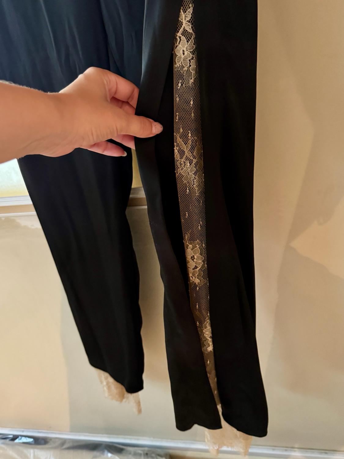 Hand showing the side detail of a pair of black trousers with a strip of sheer beige floral lace. Neutral background.
