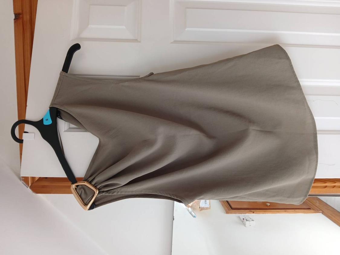 Khaki asymmetric top with gold buckle, hanging on a black hanger with the letter S on a white door in a casual setting.