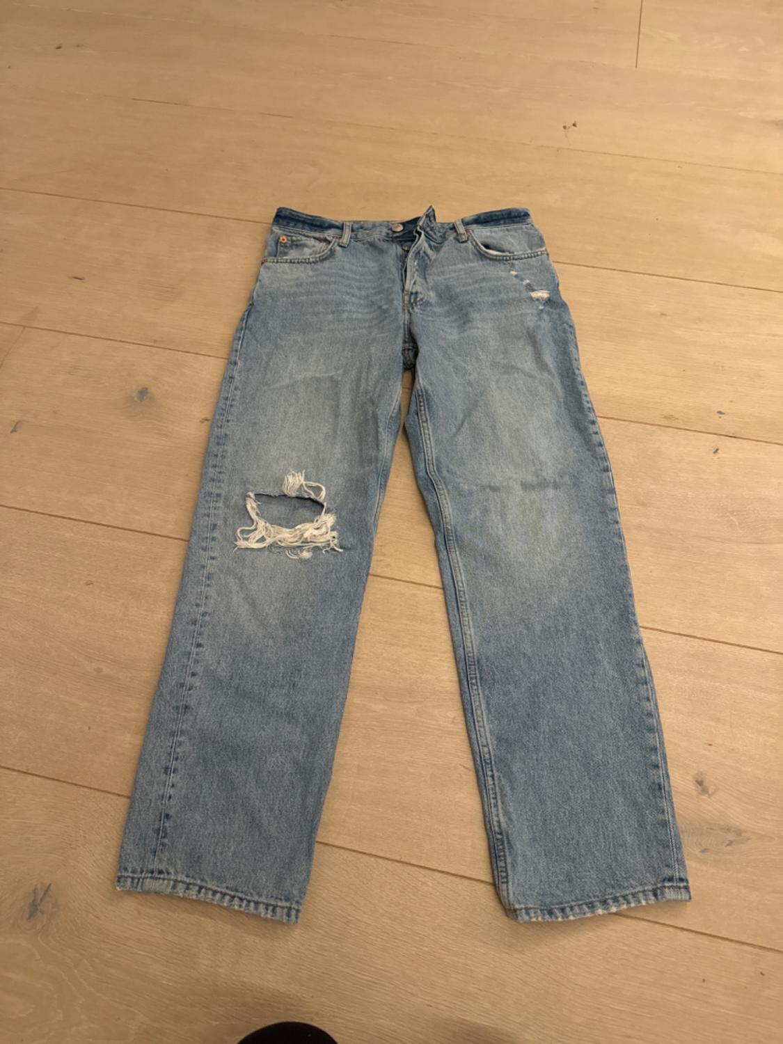 Worn blue jeans with a tear on the left leg, placed on a light wooden floor.
