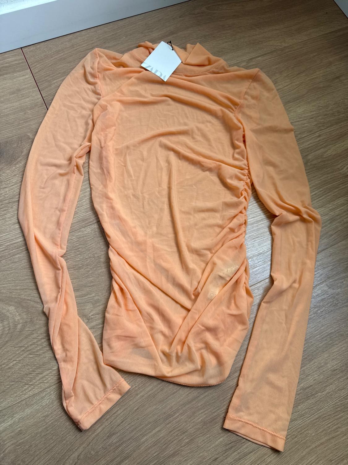 Peach-coloured long-sleeved T-shirt with white tag on wooden floor. Lightweight and wrinkled fabric.