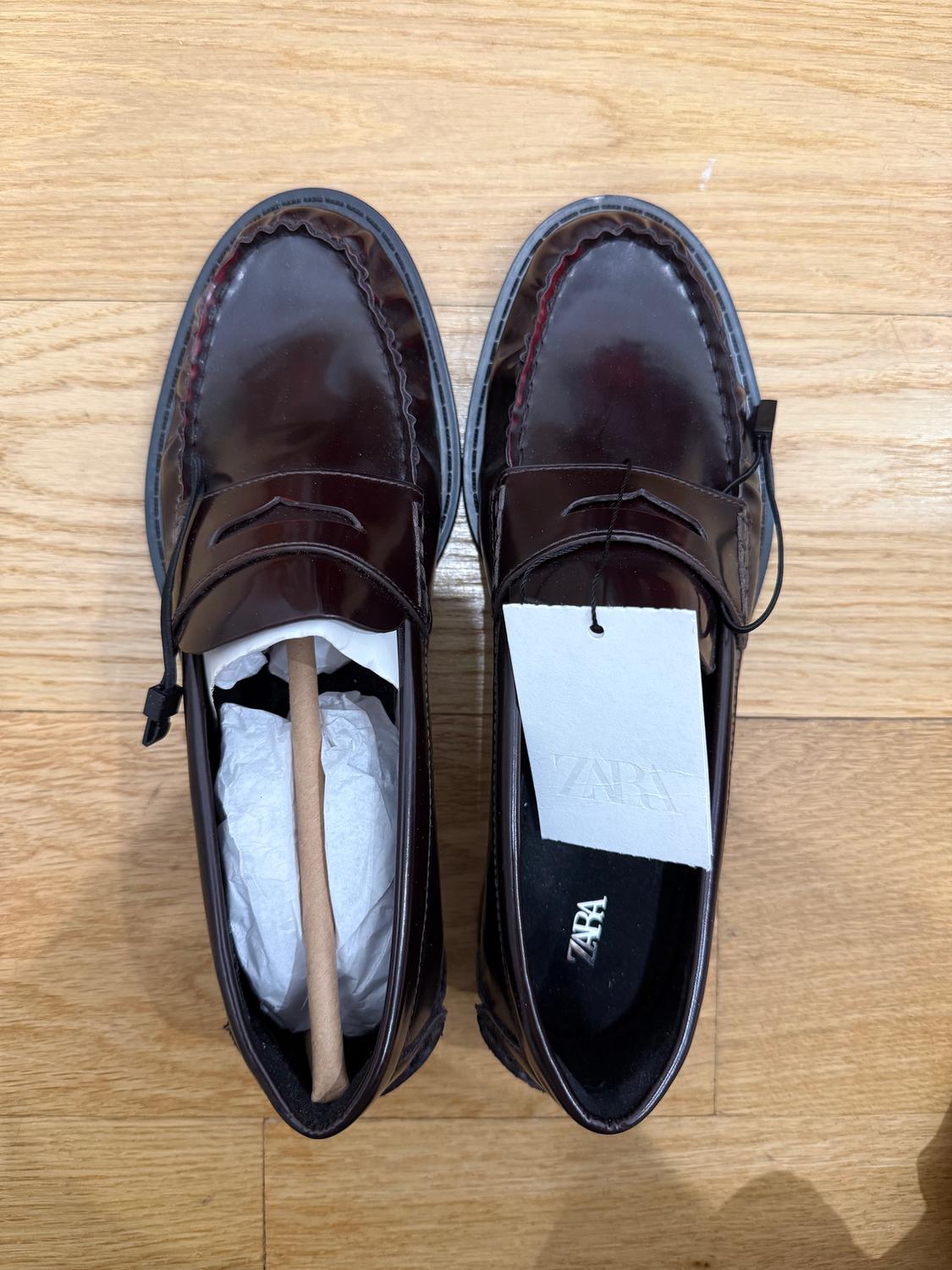 Dark brown patent leather shoes on a wooden floor, one with paper and a shoe tree, the other with a white label that says "ZARA".