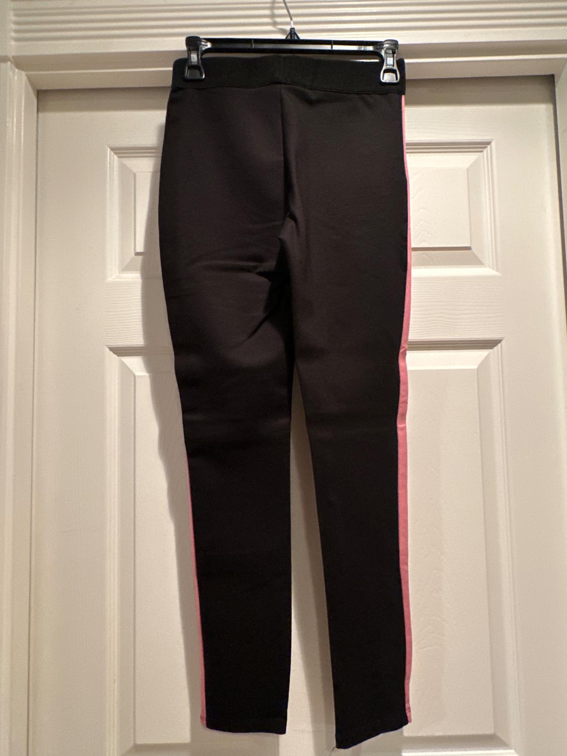 Black trousers with pink stripes down the sides, hanging on a hanger in front of a white door.