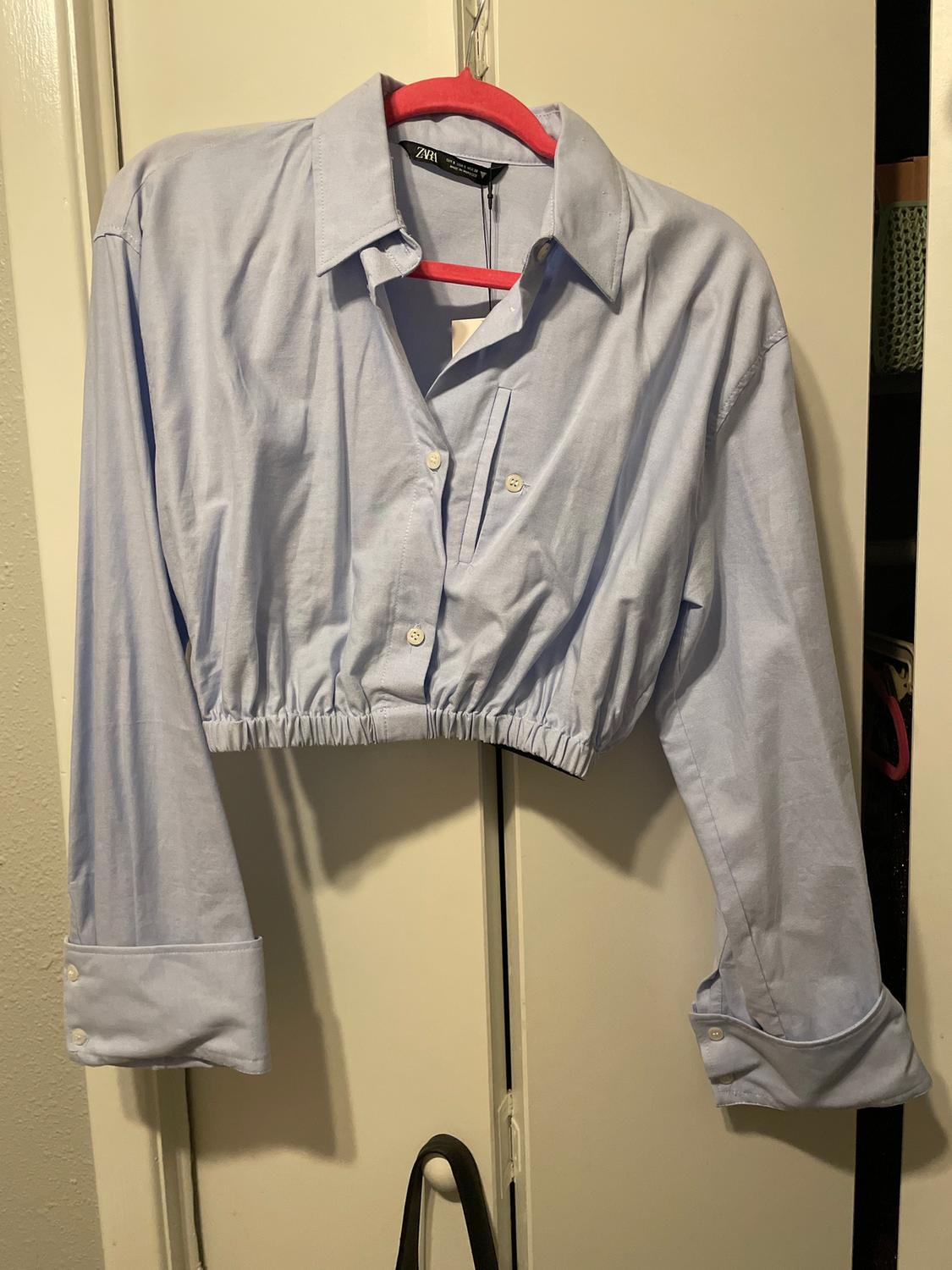 Light blue, long-sleeved shirt with an elasticated waist, hanging on a red hanger in front of a white door.