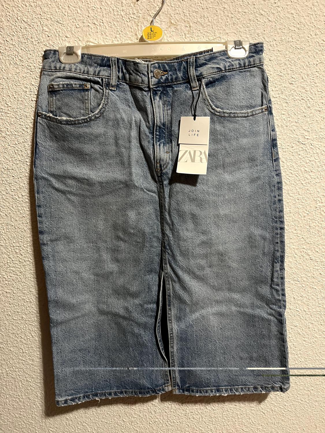 Blue denim skirt hanging on a white hanger with a "JOIN LIFE ZARA" tag, against a textured beige wall.