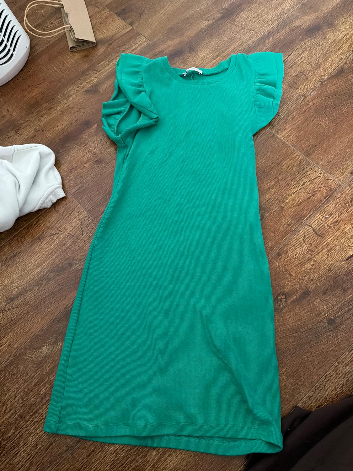 Green dress with ruffled sleeves on a wooden floor. To the left, a white sweatshirt and a paper bag.