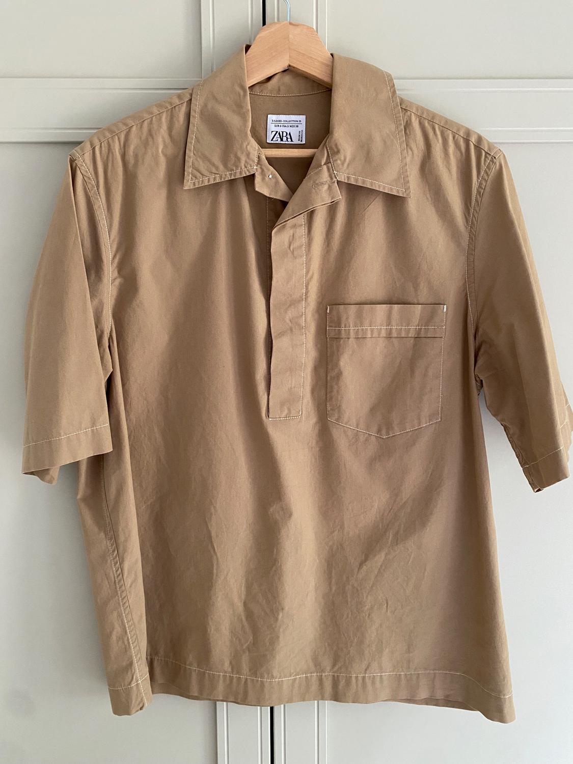 Beige short-sleeved shirt hanging on a wooden hanger. Visible label with text: "ZARA". White door background.