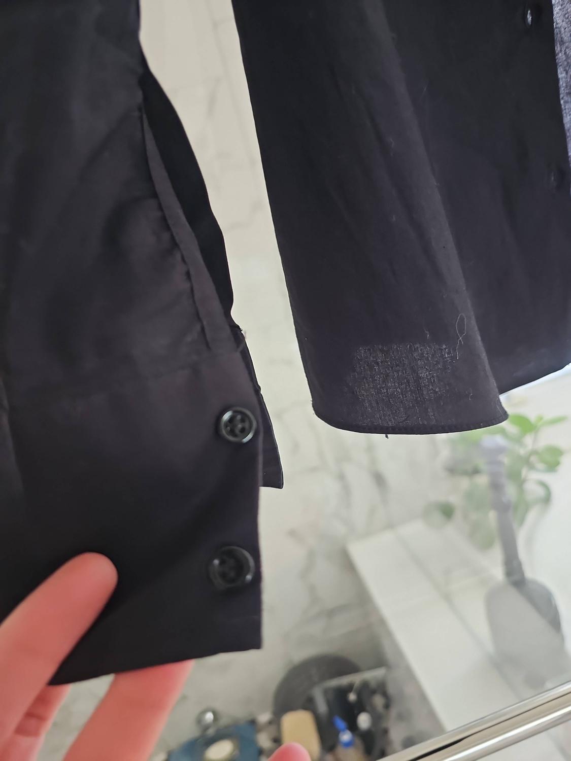 **Detailed view of the cuff of a black shirt with two buttons, held by a hand. Blurred background with a plant.**