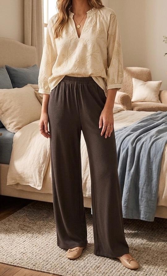 Woman in a beige blouse and wide brown trousers, standing in a cosy bedroom with a bed and cushions in neutral tones.
