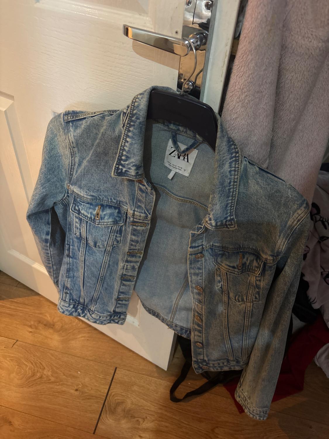 Blue denim jacket hanging on a white door. Text on label: ZARA EUR S USA S MEX 26 MADE IN BANGLADESH. Wooden floor.