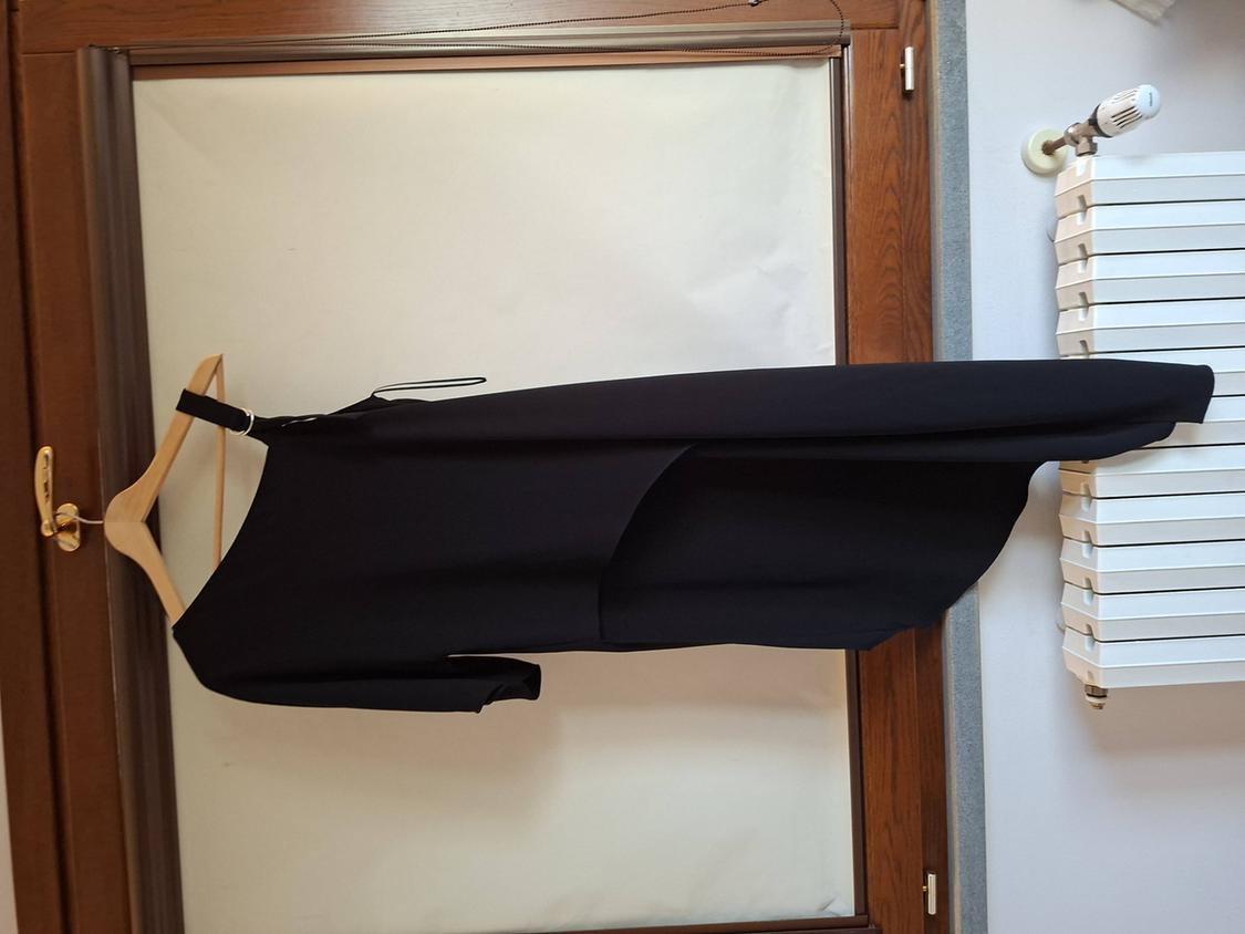 Asymmetrical black dress hanging on a wooden hanger in front of a wooden-framed window with a white radiator next to it.