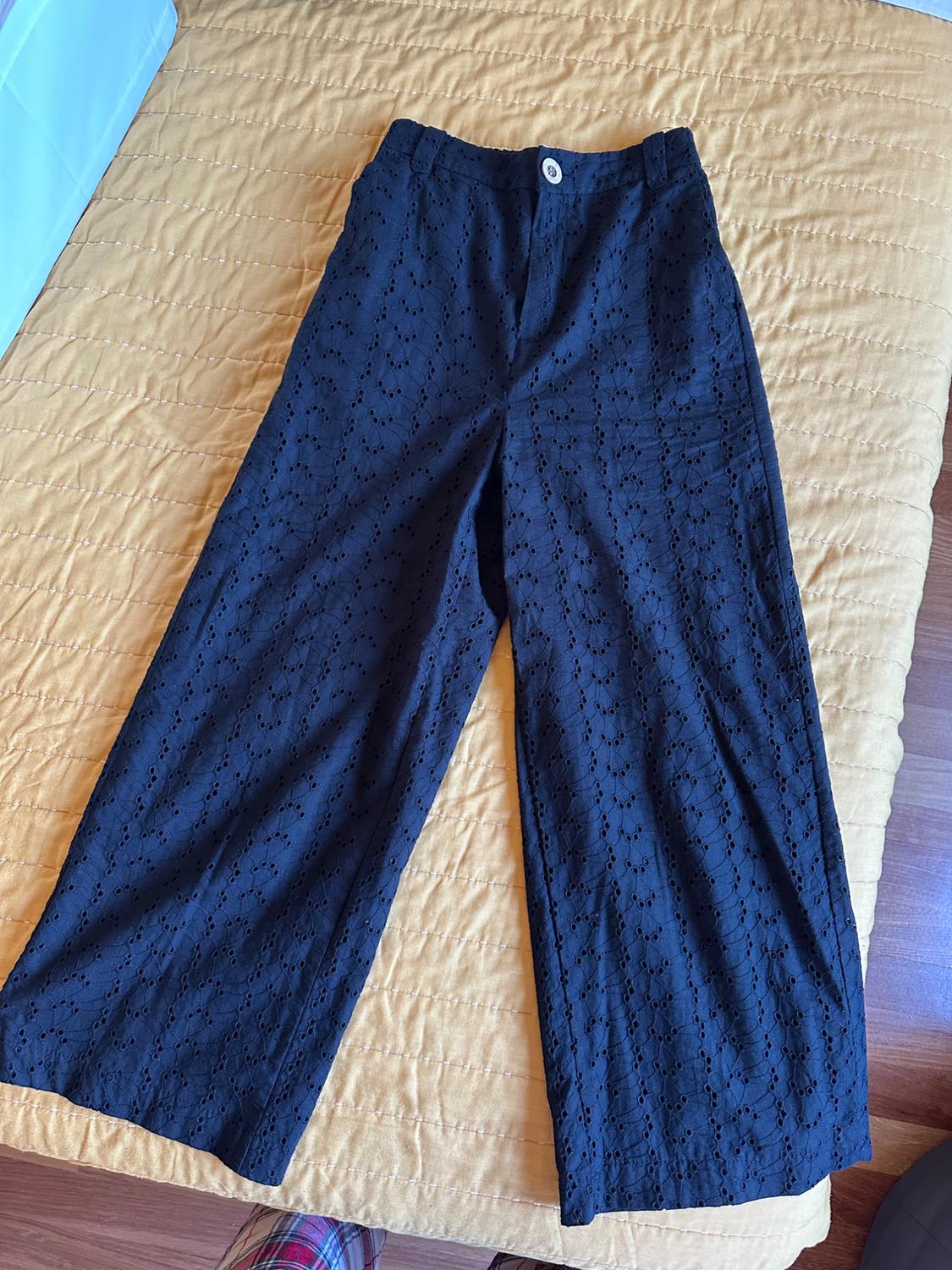 Black wide-leg trousers with broderie anglaise, laid out on a yellow quilt. Relaxed, casual style composition.