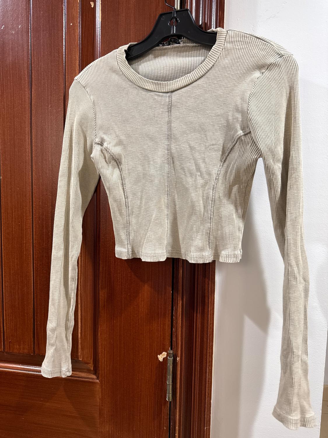 Beige, long-sleeved, slim-fit sweater hanging on a black hanger over a wooden door.