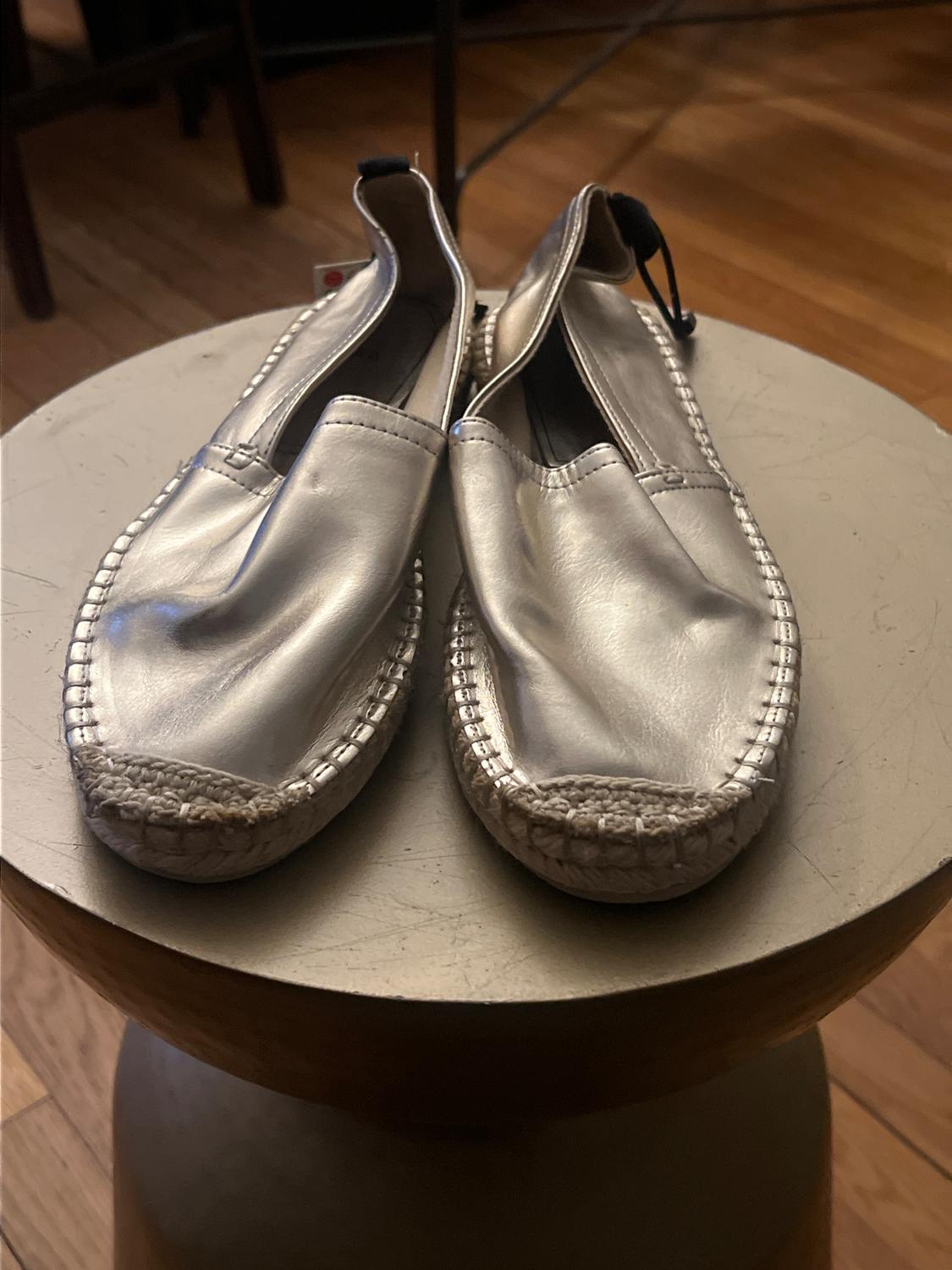Silver leather espadrille shoes, placed on a round wooden table in an indoor setting.