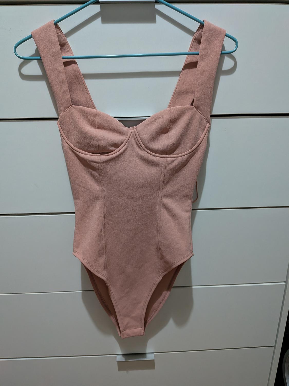 Pink bodysuit with structured cups and wide straps, hanging on a blue hanger in front of white drawers. Relaxed atmosphere.