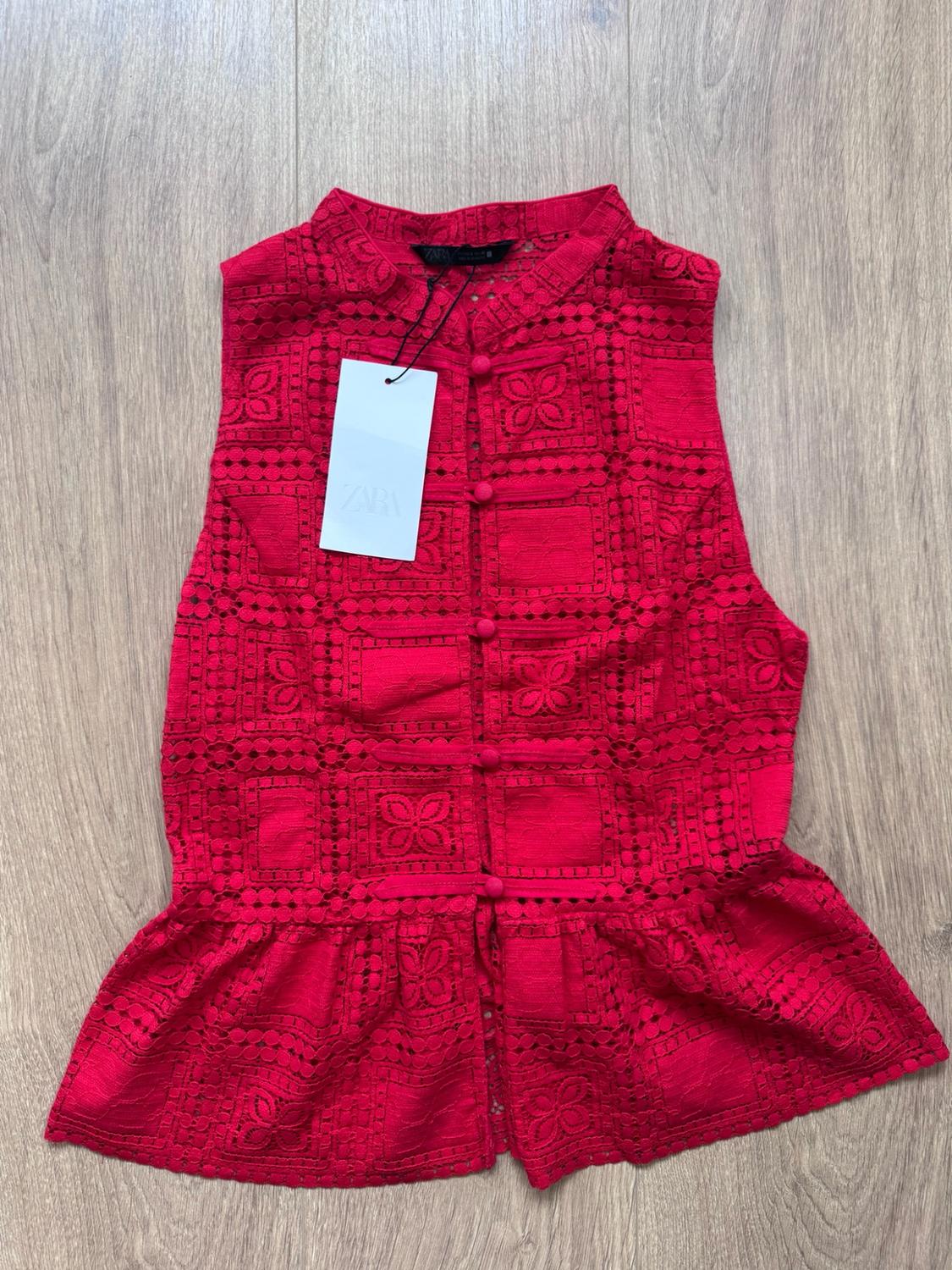 Sleeveless red top with openwork embroidery, front buttons and a flared hem on a wooden floor. Label bearing the text ZARA.