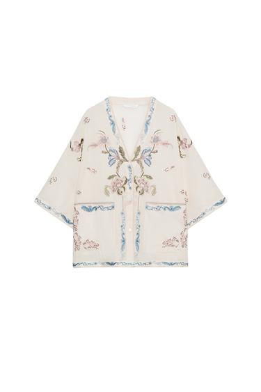 Front view of a raw shirt with a V-neck, wide sleeves, and floral embroidery in blue and pink. Patch pockets.