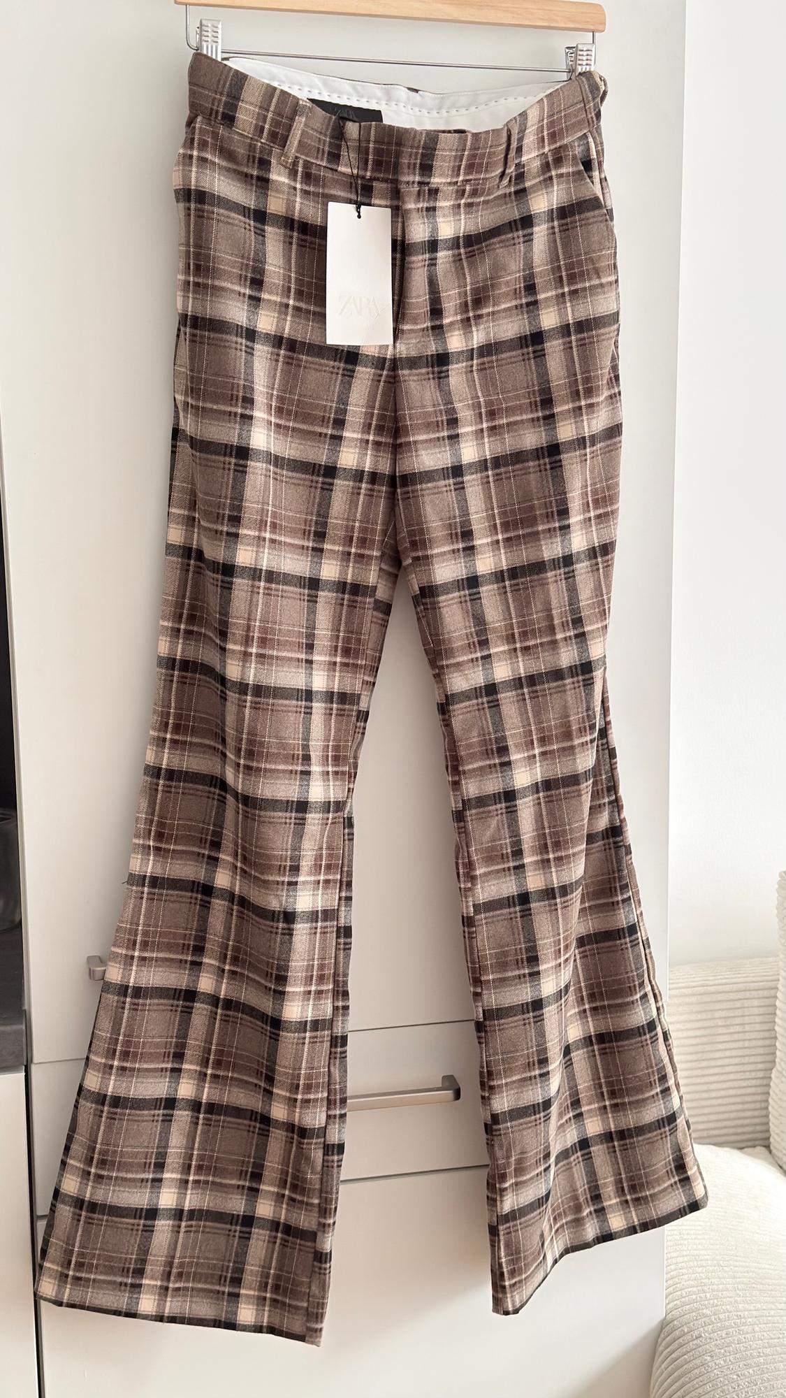 Brown checked trousers hanging on a hanger, with a visible label. White background and bright setting.