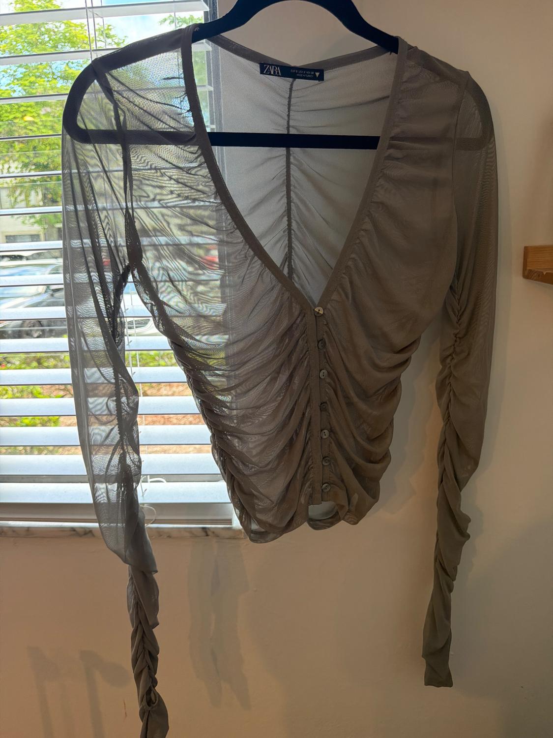 Semi-sheer gray long-sleeve top with ruching and buttons, hanging on a hanger in front of a window with blinds. ZARA label.