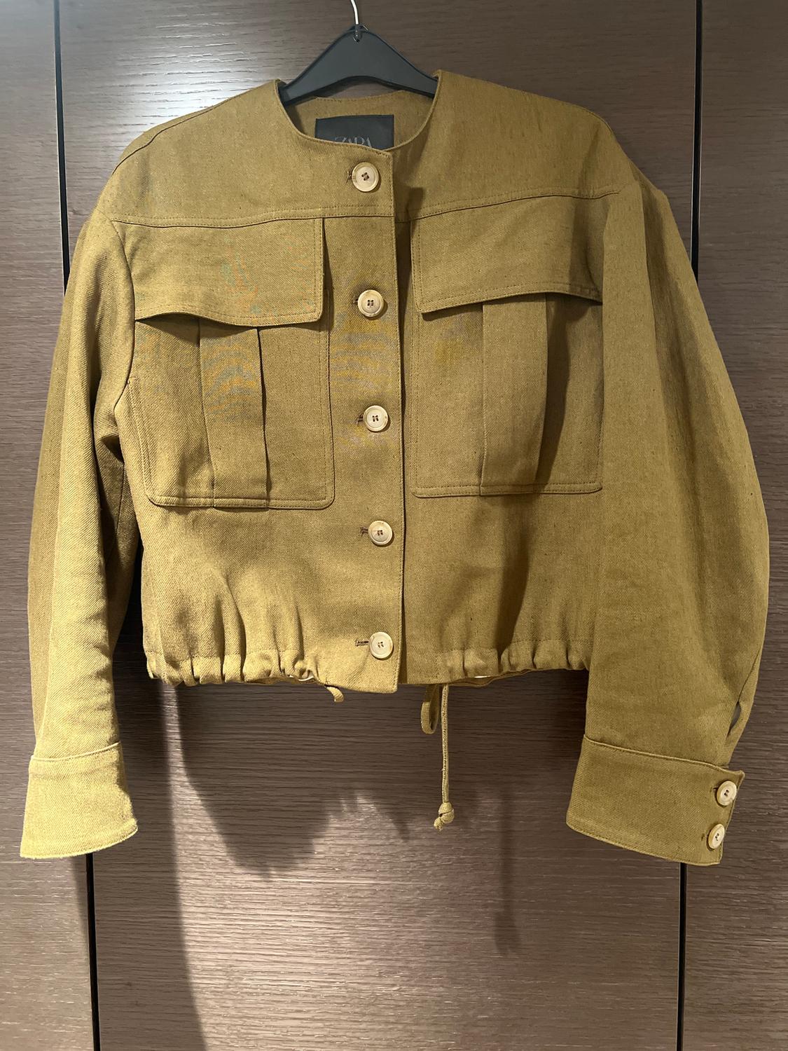 Khaki short jacket with large buttons and front pockets, hanging on a hanger against a dark wooden background.