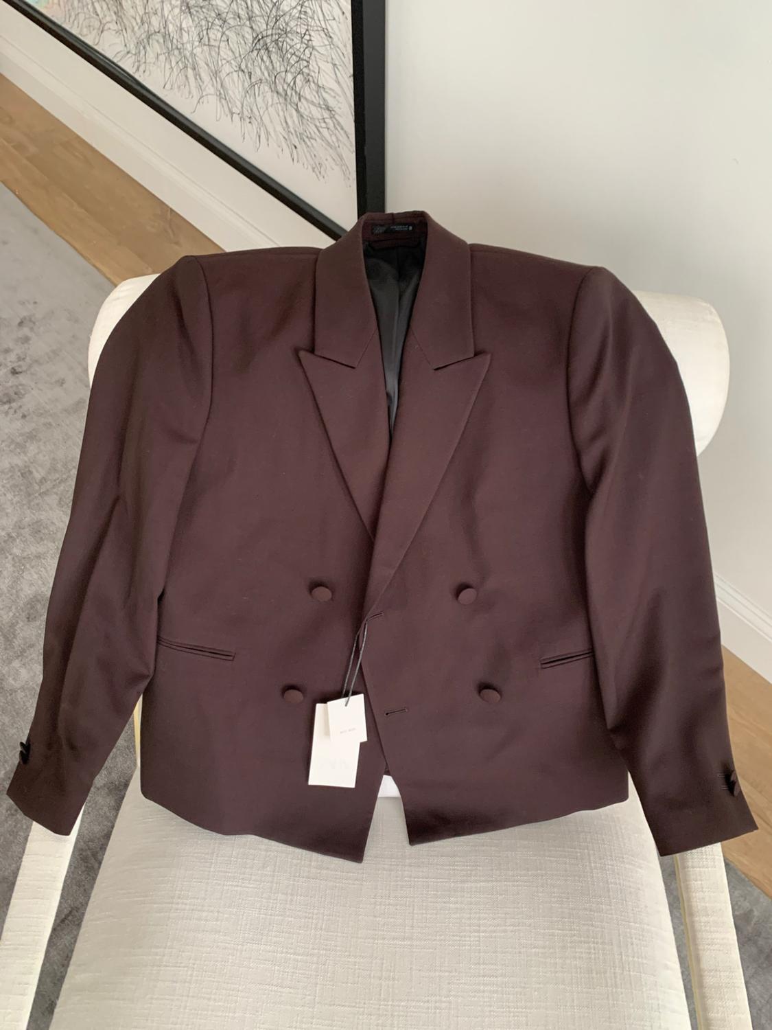 Dark brown double-breasted blazer with matching buttons and a label, placed on a white upholstered chair in a modern interior.