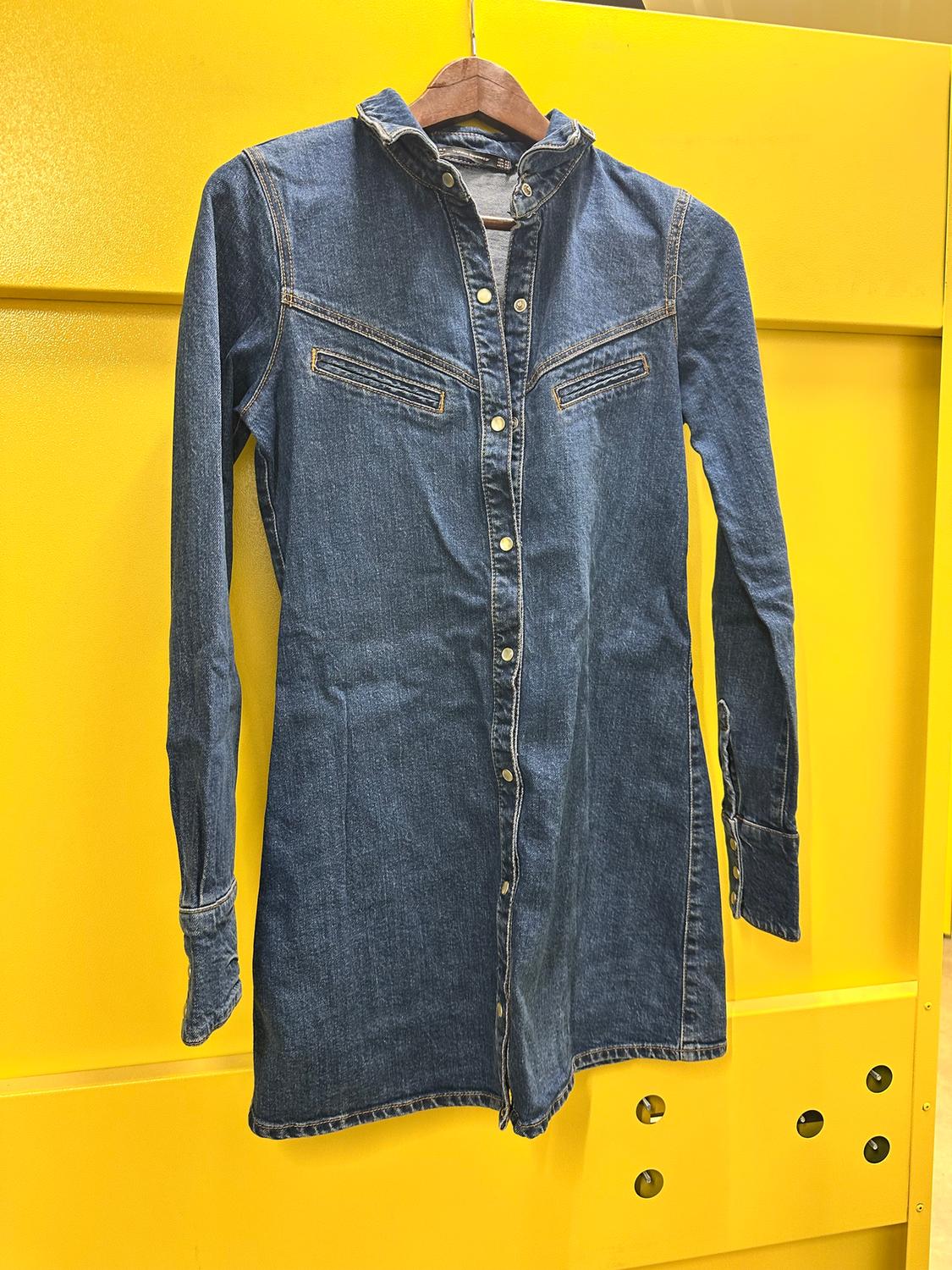 Blue denim shirt dress hanging on a wooden hanger against a bright yellow background.