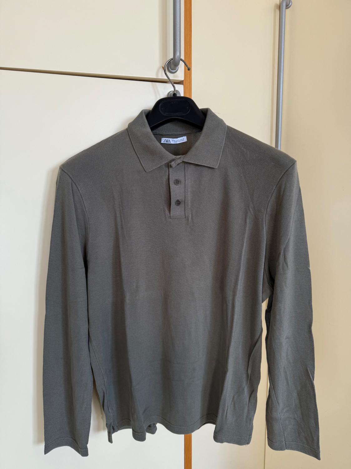 Long-sleeved grey polo shirt hanging on a black hanger, with a beige wardrobe background. Visible label on the collar.