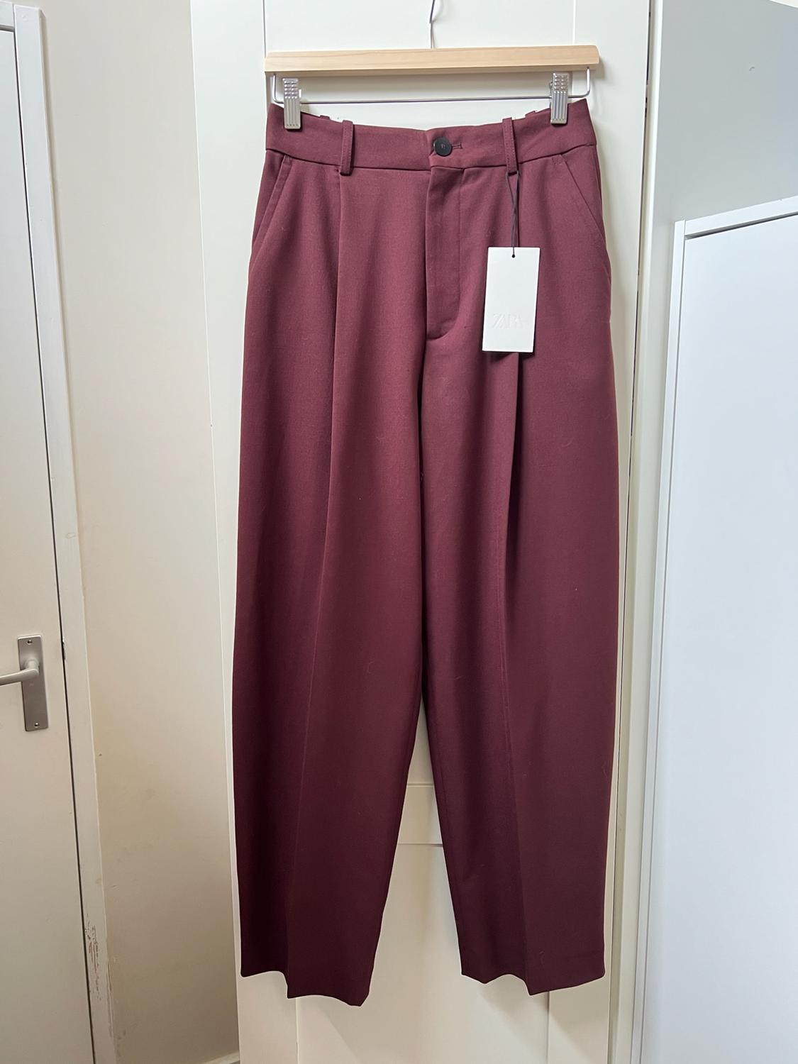 Burgundy pleated trousers hanging on a wooden hanger in front of a white wardrobe. It has a label with the text ZARA.