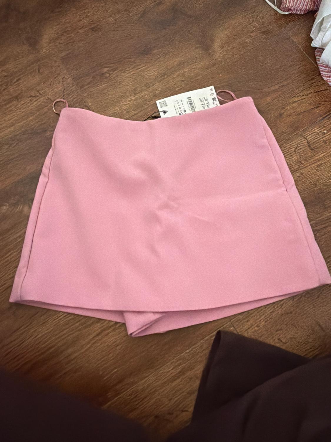Pink short skirt on a wooden floor, with a visible label at the top. Relaxed and casual atmosphere.