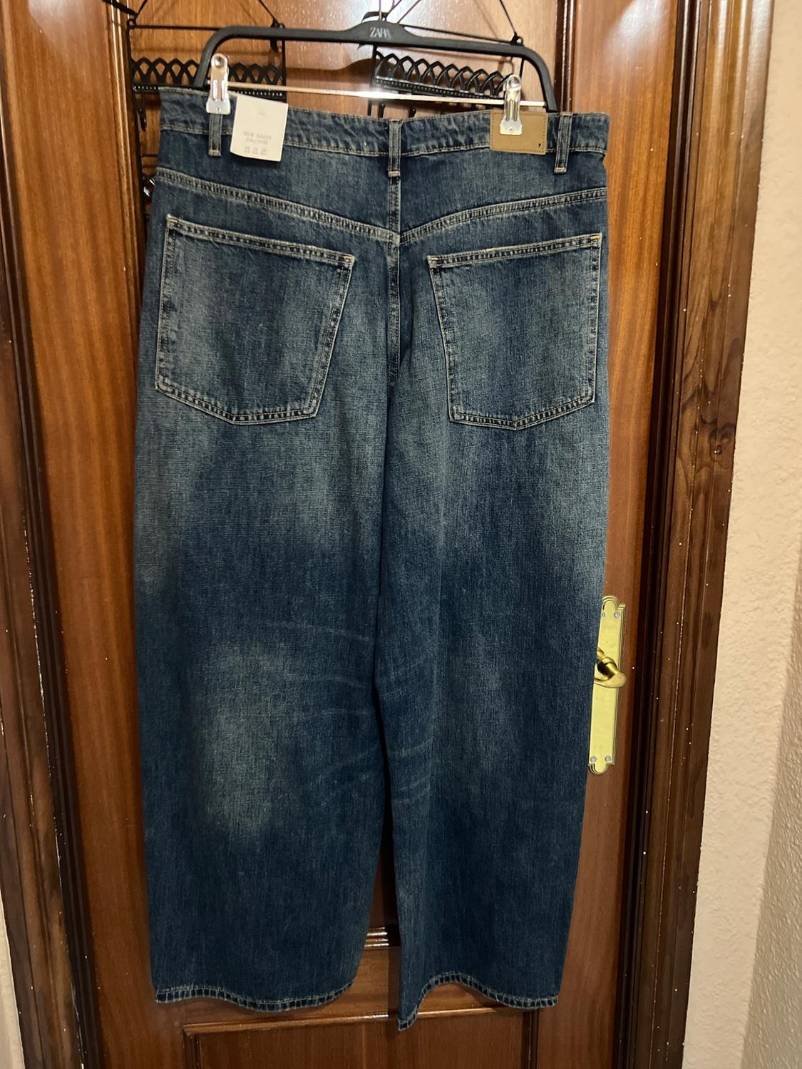 Blue jeans seen from behind, hanging on a wooden door. Text: ZARA on the hanger and NEW BAGGY BALLOON on the label.