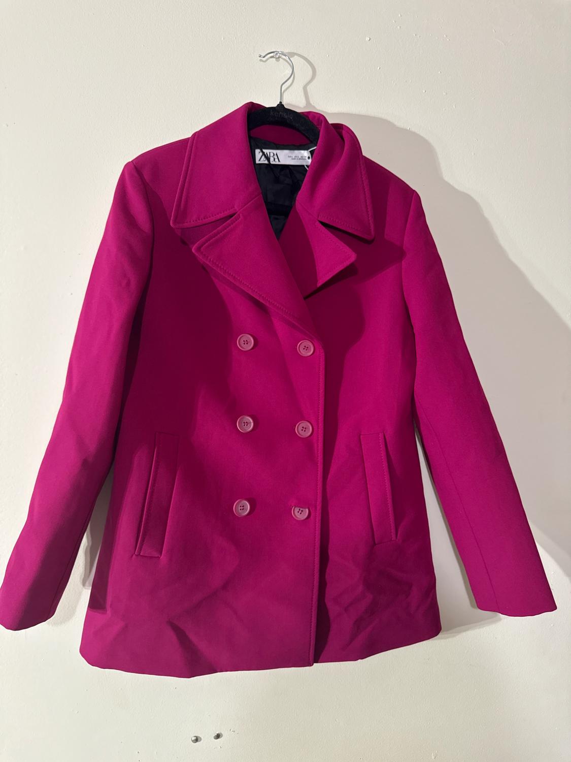 Fuchsia double-breasted coat hanging on a hanger against a white wall. Label visible on the inside of the neck.