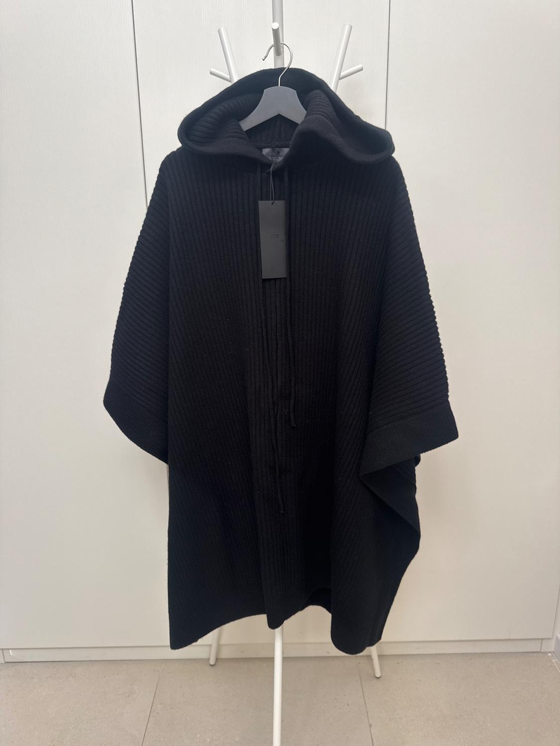 Black chunky knit cape with a hood, hanging on a white hanger against a light background. Black label visible on the front.