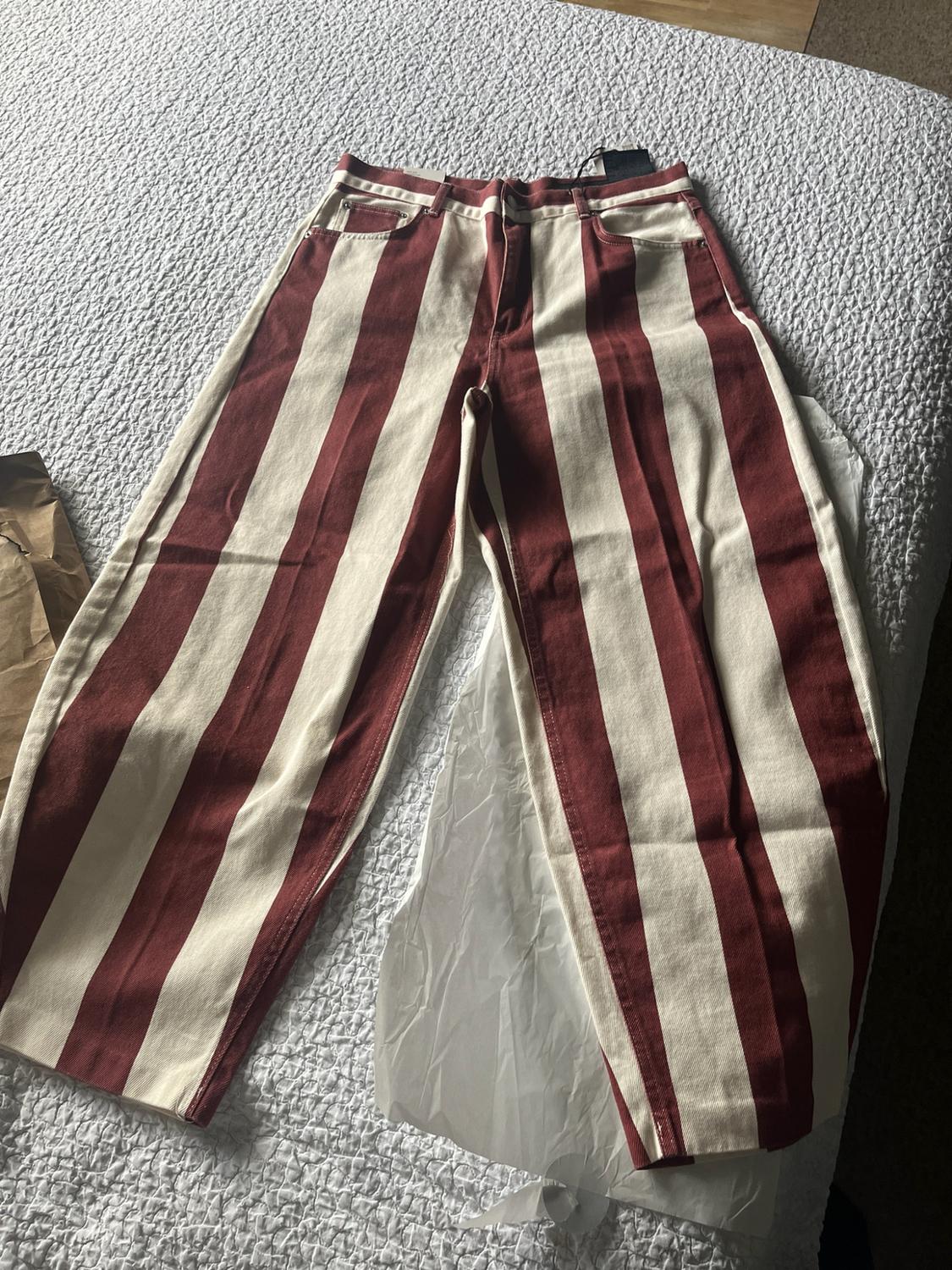 Wide-leg trousers with vertical maroon and beige stripes, laid out on a bed with a white bedspread and tissue paper, creating a casual atmosphere.