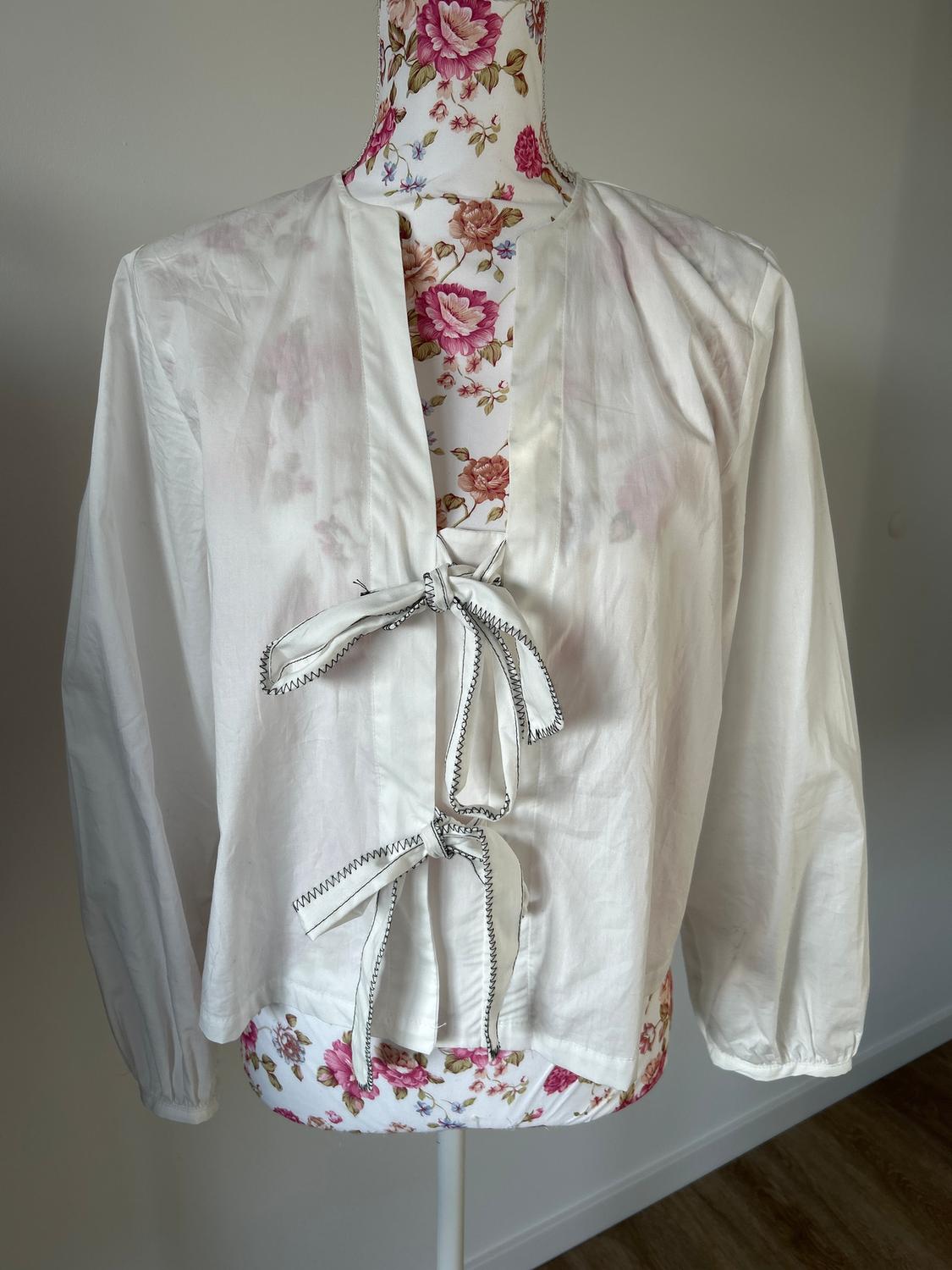 Long-sleeved white blouse with a bow at the front, displayed on a mannequin with a floral print against a neutral background.