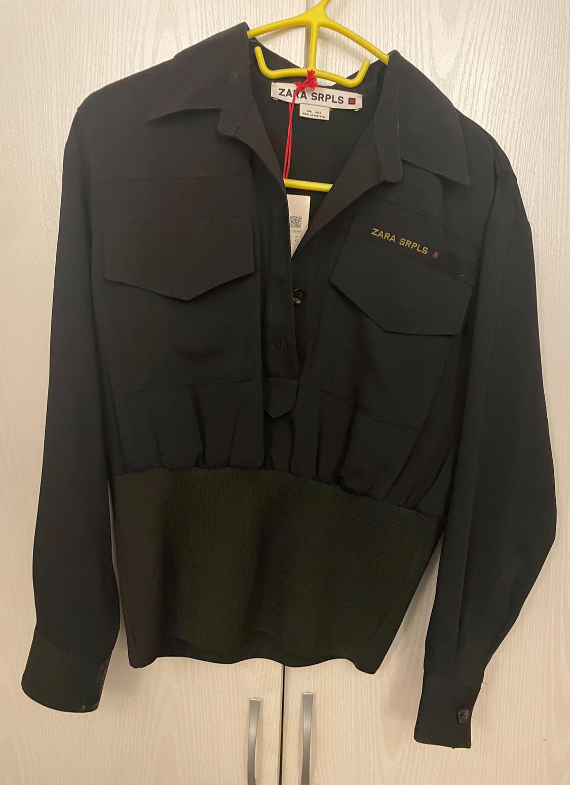 Black long-sleeved shirt with chest pockets, hanging on a yellow hanger. Visible text: "ZARA SRPLS".