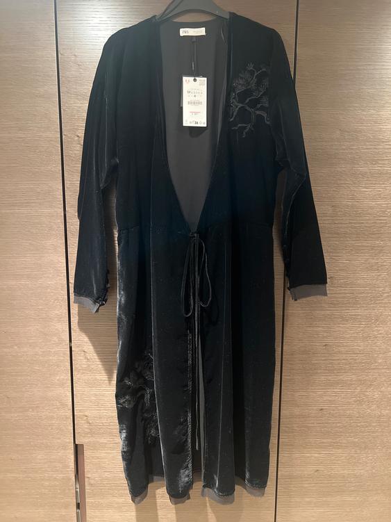 Black velvet dress with floral embroidery on the shoulder, hanging on a wooden hanger. Visible label on the collar.